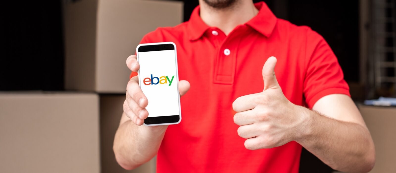 partial view of cheerful delivery man with smartphone with ebay logo on screen showing thumb up